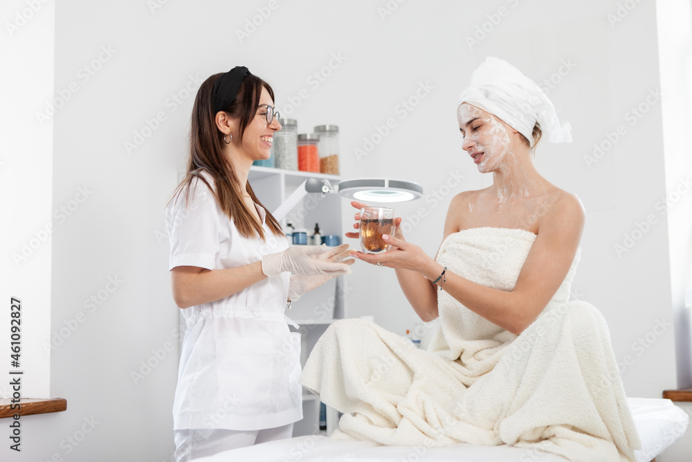 Woman beautician treats her client with cup of tea. Beauty woman in beauty salon. Facial skin care Woman beautician treats her client with cup of tea. Beauty woman in beauty salon. Facial skin care