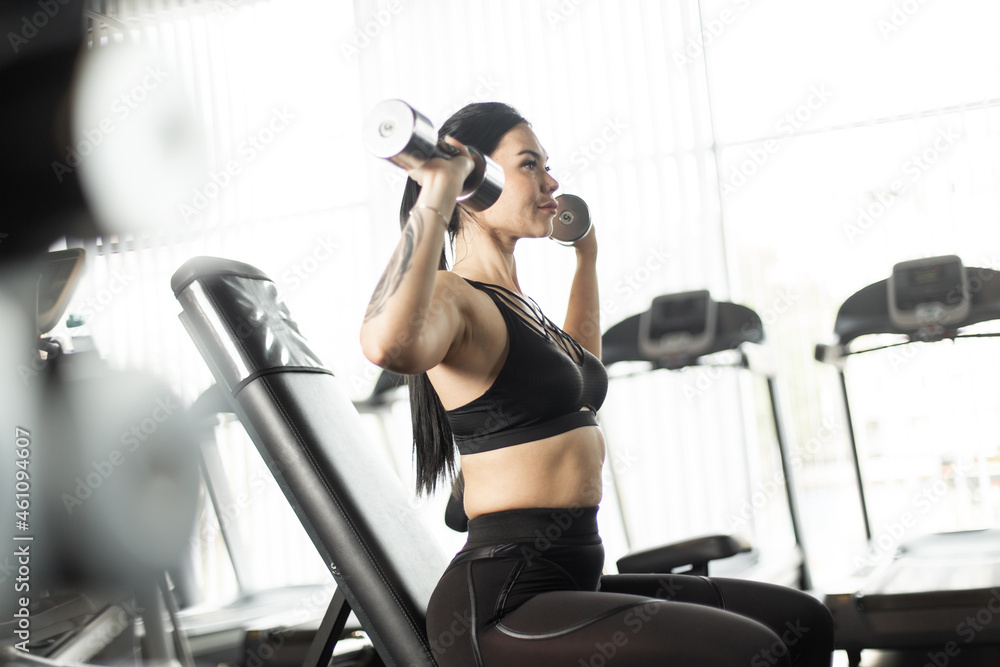 Young fitness brunette woman doing overhead dumbbell press while sitting on bench in the gym Young fitness brunette woman doing overhead dumbbell press while sitting on bench in the gym