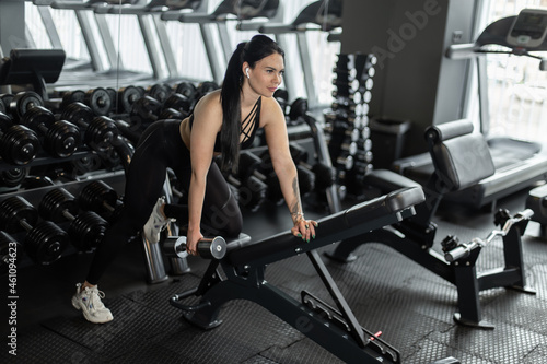 Sexy fitness brunette woman doing dumbbell rows with one hand leaning on a bench in the gym