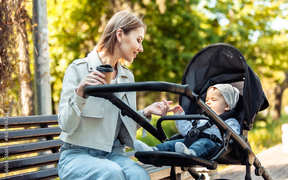 Obraz premium Beautiful Caucasian mother sits on a bench with baby in stroller outdoor