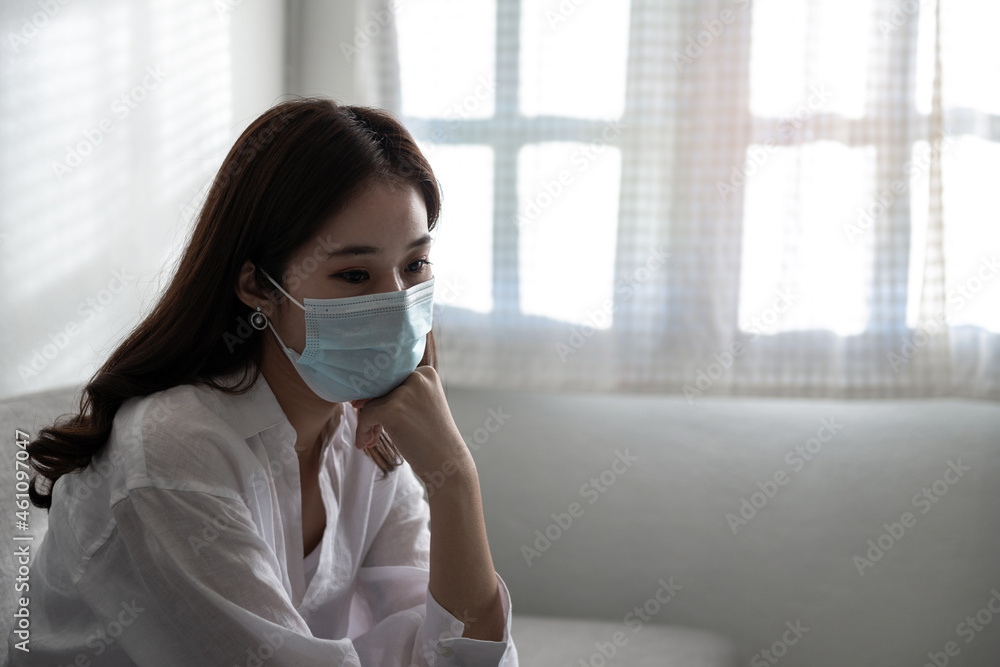 Close-up of a sad Asian patient woman wearing a face mask at home or hospital alone on a sofa waiting for a doctor and vaccine. Health care during Coronavirus or Covid-19 quarantine concept Close-up of a sad Asian patient woman wearing a face mask at home or hospital alone on a sofa waiting for a doctor and vaccine. Health care during Coronavirus or Covid-19 quarantine concept