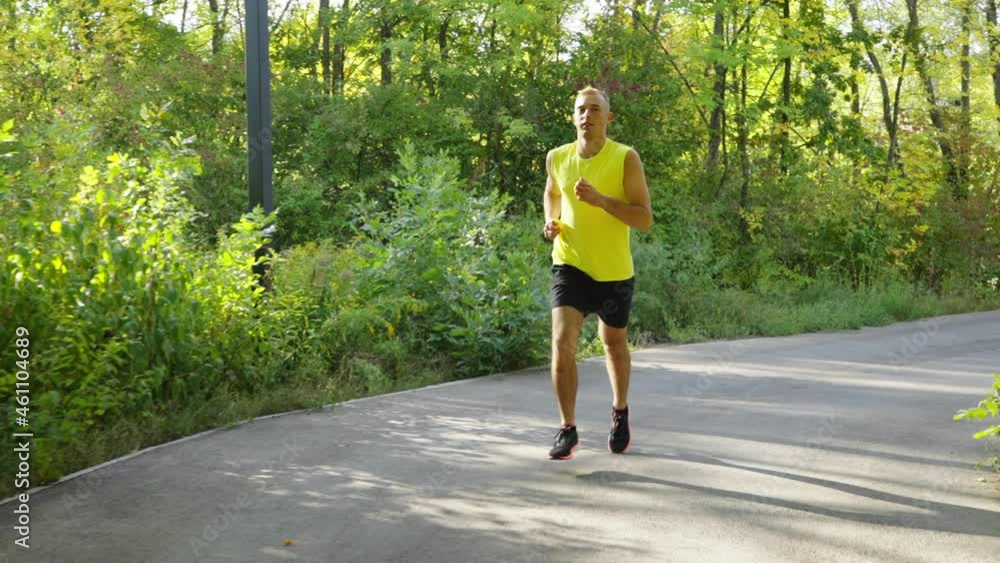 Athletic man wearing yellow shirt running in park in slow motion. Tracking shot of runner training on sunny morning. Concept of healthy lifestyle Athletic man wearing yellow shirt running in park in slow motion. Tracking shot of runner training on sunny morning. Concept of healthy lifestyle