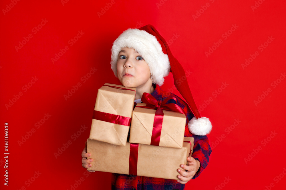 Happy holiday and Merry Christmas. Portrait of a boy in a cap with gifts in his hands on a red background. A place for text. Happy holiday and Merry Christmas. Portrait of a boy in a cap with gifts in his hands on a red background. A place for text.