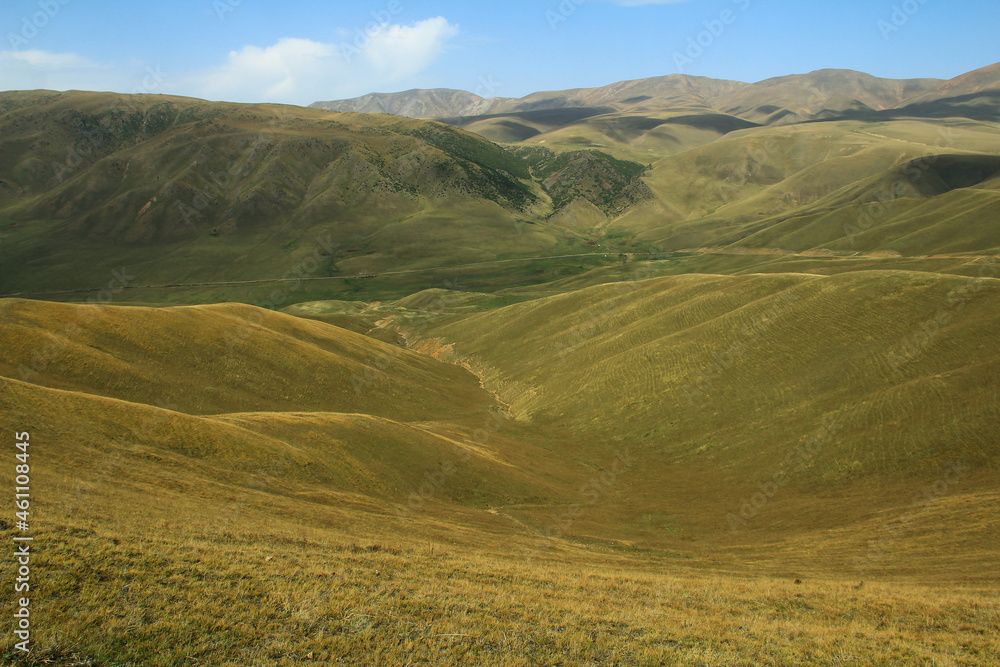 Top view of a wide mountain grassy valley on the Assy plateau, in front of the soft hills, in the distance mountain ranges, the sky with small clouds, summer, sunny