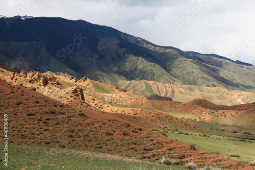 Clay red mountains with relief ledges on the Assy plateau, in the background a large mountain with ridges covered with forest, sky with clouds, sunlight, summer