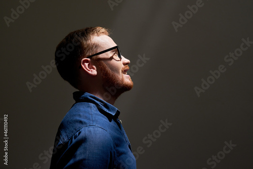 Side view of smiling handsome young red-bearded man in eyeglasses looking at light in dark room, ambitions concept