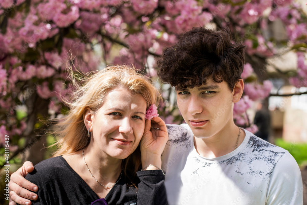 A teenage boy with a manly look hugs his mother on a background of spring pink flowers. The woman is happy and smiling - she has a good relationship with her teenage son A teenage boy with a manly look hugs his mother on a background of spring pink flowers. The woman is happy and smiling - she has a good relationship with her teenage son