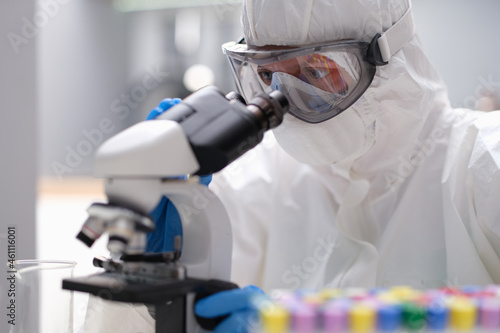 Scientist chemist in protective anti-plague suit looking through microscope in laboratory