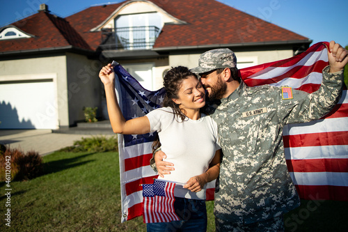 Portrait of husband soldier in uniform on military leave kissing his lovely wife and holding American flag in front of their house. photo