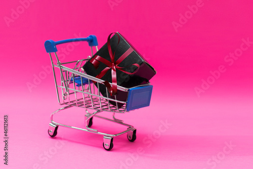Creative concept with shopping trolley with gifts isolated on pink background. Gifts wrapped in kraft paper with a red ribbon and bow.