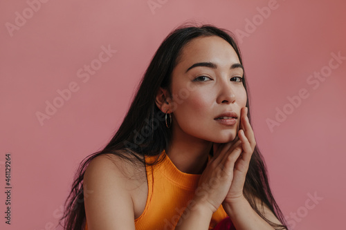 Close-up studio portrait of young asian woman mysteriously looking at camera on pink background. Long-haired brunette in orange top touches her cheek with fingers. © Look!
