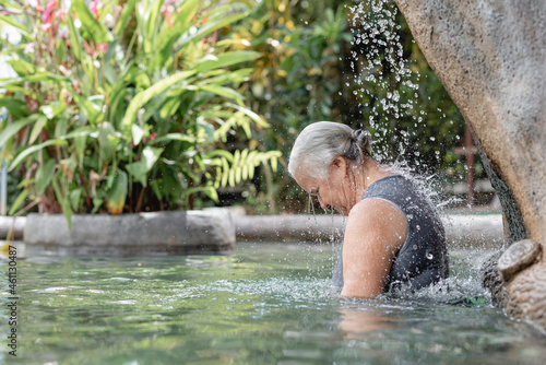 Mujer adulta mayor con cabello canoso en una piscina de agua termal disfrutando mientras cae agua sobre su espalda photo