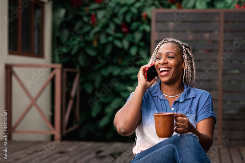 Mujer madura afroamericana con cabello con trenzas sonriendo mientras habla por celular y toma una taza de café o té photo
