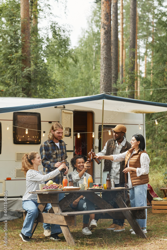 Vertical full length view at diverse group of young people clinking beer bottles while enjoying picnic outdoors at campsite with trailer van Vertical full length view at diverse group of young people clinking beer bottles while enjoying picnic outdoors at campsite with trailer van