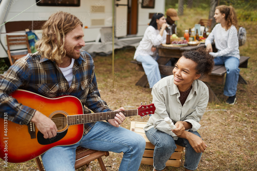 Portrait of young couple playing guitar while camping with friends in forest, copy space