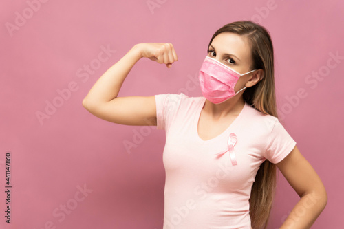 Woman shows strength as symbol of fighting breast cancer in front of pink background and wearing face mask High quality photo