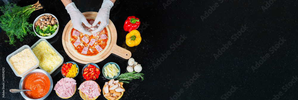 Top view of chef adding ham into the dough of a homemade pizza with tomato sauce while wearing gloves and pizza ingredients, spices on black background. Pizza menu. View from above. Space for text.