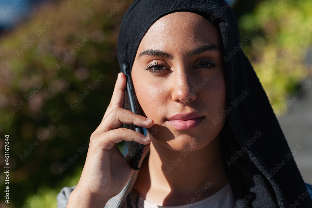 Close up view of a muslim woman talking with her phone while looking at camera in the park Close up view of a muslim woman talking with her phone while looking at camera in the park