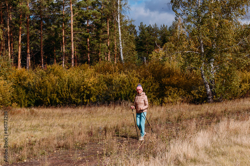 elderly sixty-year-old woman in sportswear is engaged in Nordic walking with sticks in the forest in the autumn © Alyona