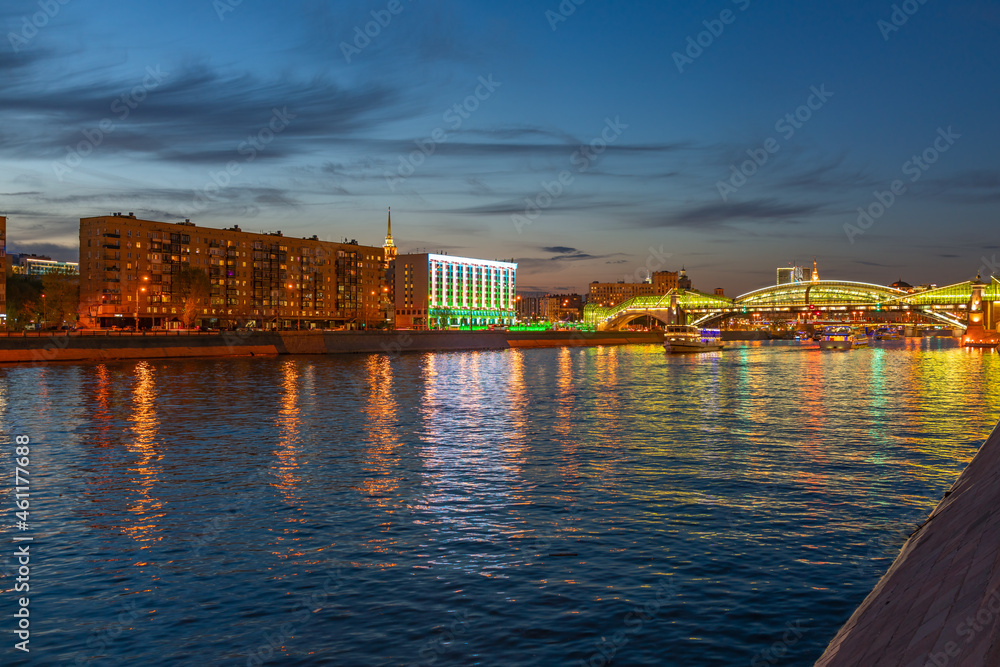 View of the colorful Bogdan Khmelnitsky bridge illuminated at night reflecting in the Moskova river. Moscow Kiyevsky railway station at night. Moscow, Russia