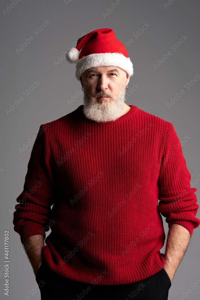 Stylish Santa. Middle aged man with beard wearing Christmas hat and red sweater looking at camera while standing isolated over gray background