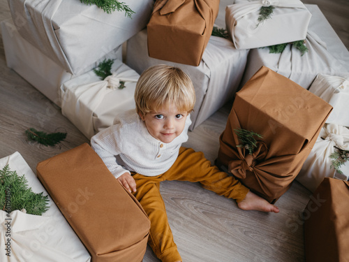 Two years old caucasian blond child dressed in white and mustard sitting on a wooden floor looking at the camera surrounded by Christmas presents wrapped in furoshiki technique