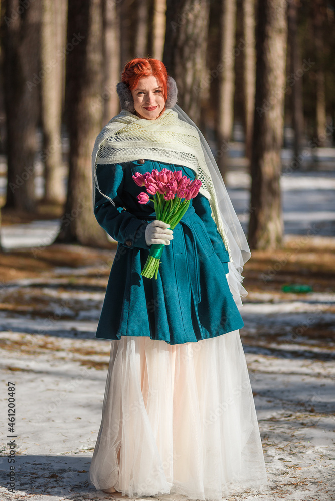 beautiful girl in a green coat and a wedding dress with a bouquet of flowers in the forest in winter