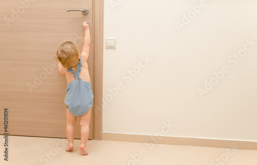Blonde caucasian baby girl of 1-2 years old trying to open locked wooden brown door with key indoors. Curious, funny kid.Child safety idea.Copy space for text. Beige neutral colors