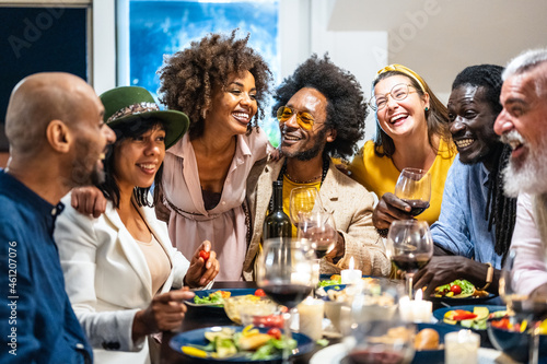 Different people of age and ethnicity eating a vegan dinner. Multiethnic group of friends having fun while sharing a meal in a warm and welcoming house  adults drinking red wine and laughing
