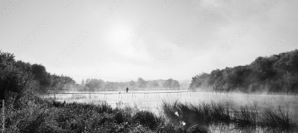 Fototapeta premium Small bridge across beautiful river in misty morning with silhouette of lone fisherman. Black and white image.