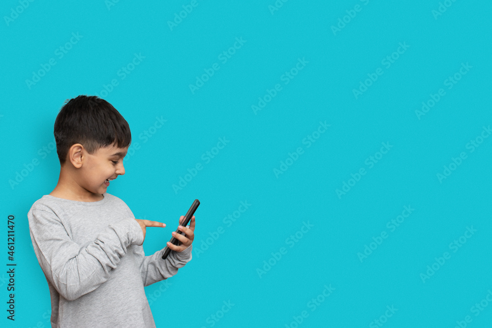 Excited boy smiling and poiting at screen on smartphone. on plain blue background with copy space Excited boy smiling and poiting at screen on smartphone. on plain blue background with copy space