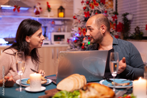 Joyful couple watching entertainment movie on laptop computer while sitting at dining table in xmas decorated kitchen. Happy family celebrating christmas holiday enjoying winter season together