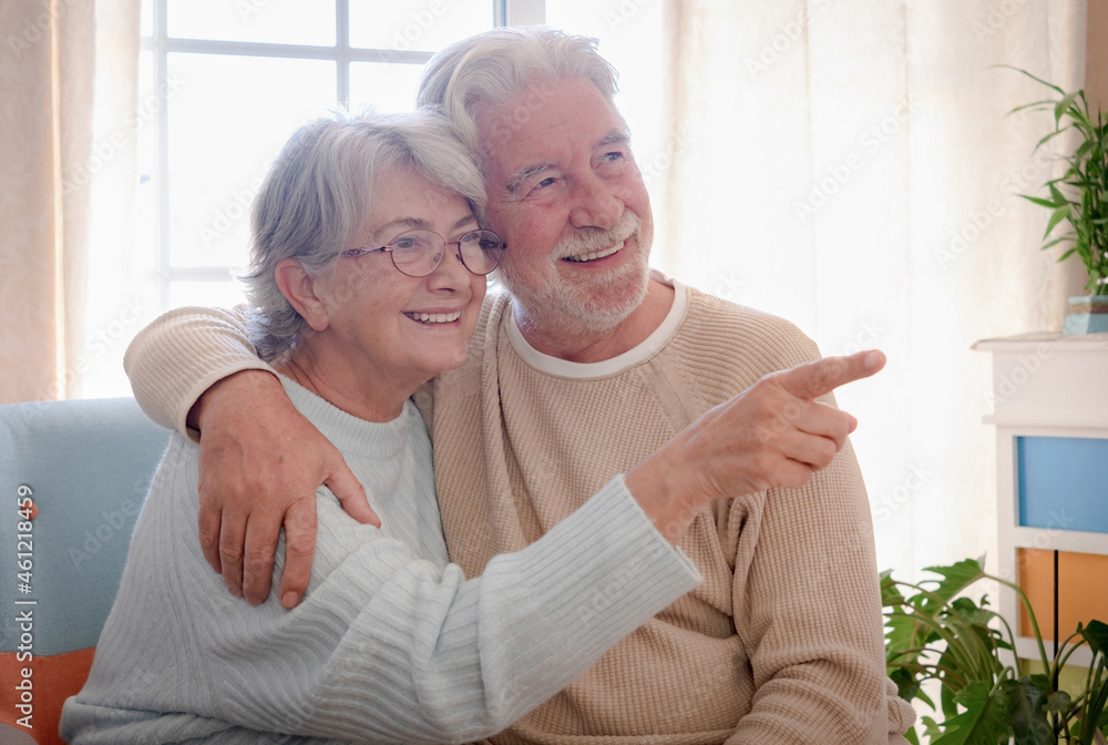 Smiling senior couple sitting at home hugging. Woman wearing eyeglasses indicating with a finger. bright light from window Smiling senior couple sitting at home hugging. Woman wearing eyeglasses indicating with a finger. bright light from window