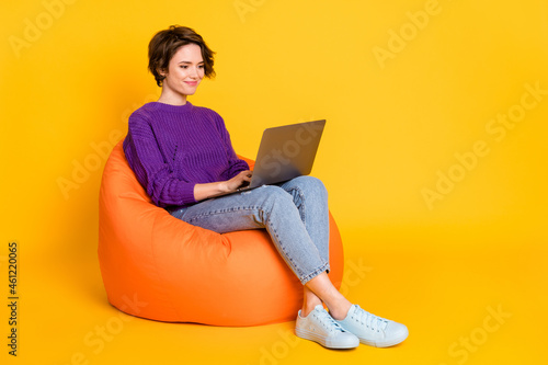 Portrait of attractive focused cheerful girl sitting in chair using laptop writing e-mail isolated over bright yellow color background photo