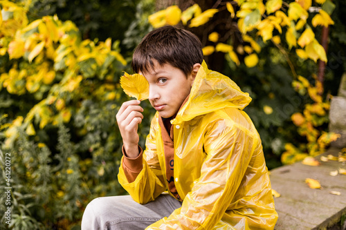 Photo of teenage boy in yellow raincoat among autumn leaves  closes one eye with leaf  looks into camera