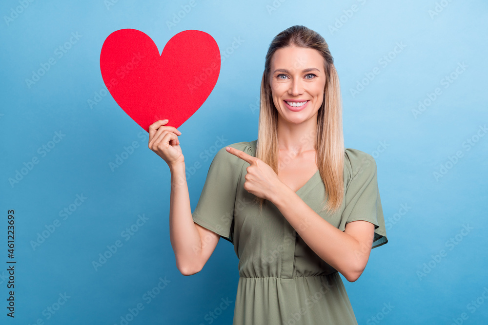 Photo of sweet shiny young woman dressed green clothes pointing finger large red heart card empty space smiling isolated blue color background Photo of sweet shiny young woman dressed green clothes pointing finger large red heart card empty space smiling isolated blue color background