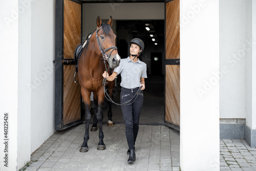 Female horseman going with her brown Thoroughbred horse near stable. Concept of animal care. Rural rest and leisure. Idea of green tourism. Young european woman wearing helmet and uniform