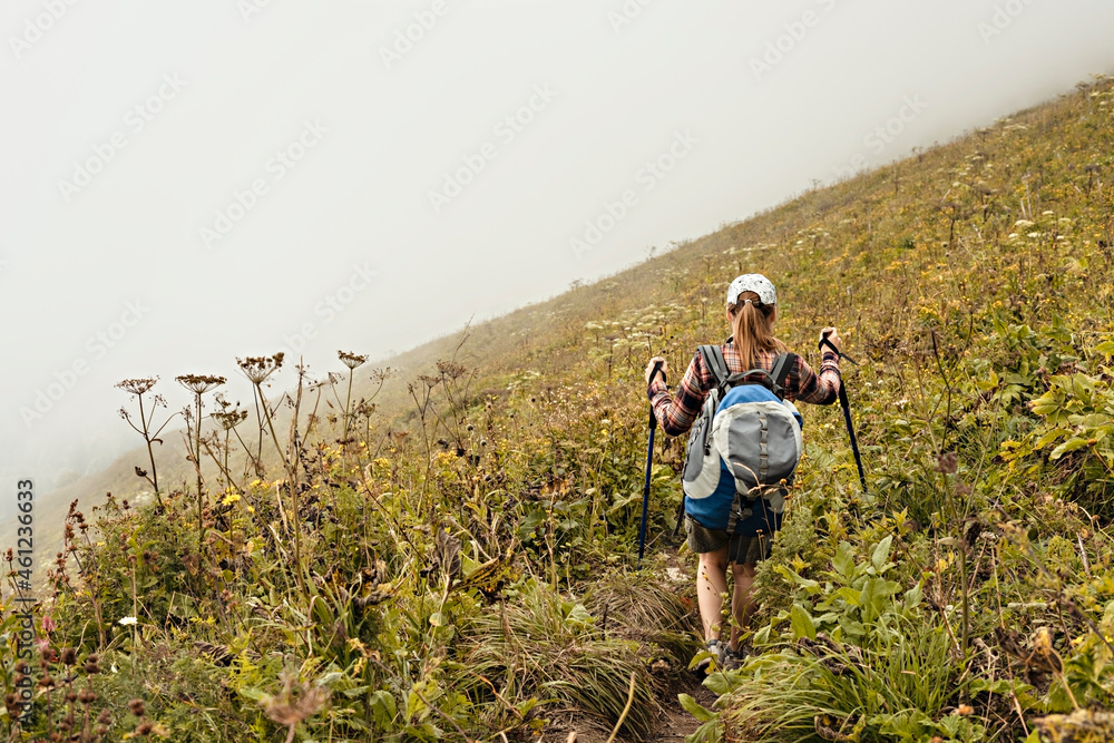rear view of young woman in plaid shirt with big backpack with trekking poles walking along mountain trail hiking in fog, healthy active lifestyle