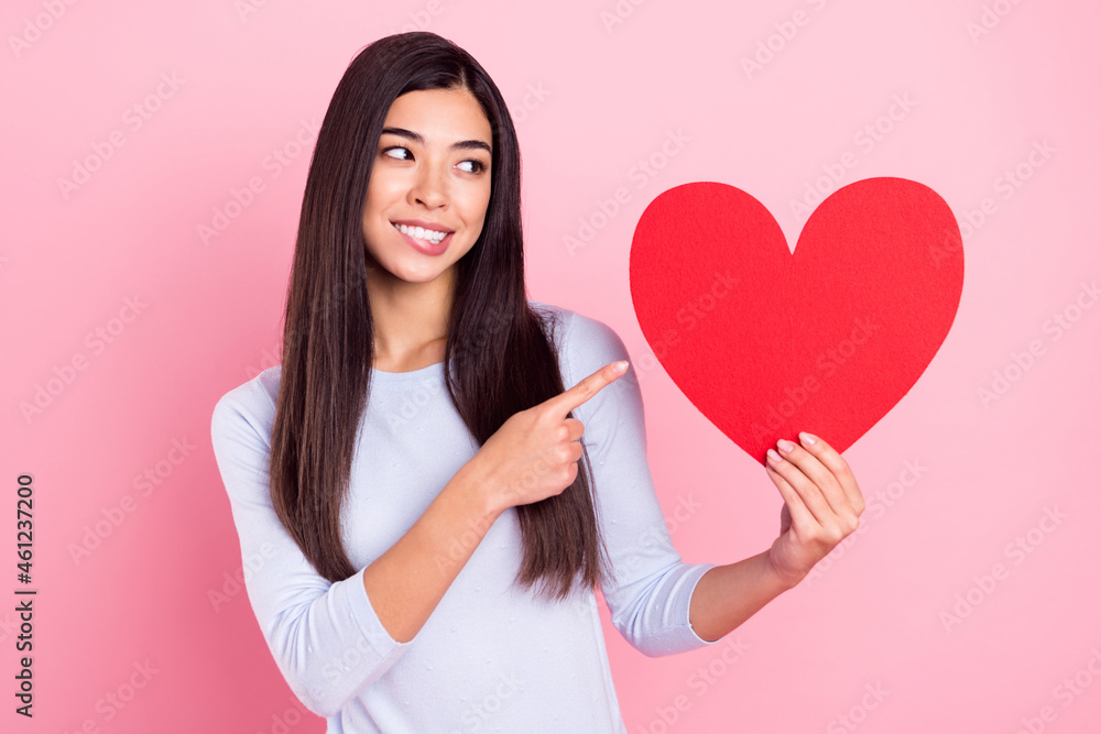 Photo portrait of girl showing finger recommending red heart fell in love biting lip isolated on pastel pink color background Photo portrait of girl showing finger recommending red heart fell in love biting lip isolated on pastel pink color background