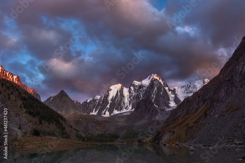 Darkness mountain landscape with great snowy mountain lit by dawn sun among dark clouds. Awesome alpine scenery with high mountain pinnacle at sunset or at sunrise. Big glacier on top in orange light.