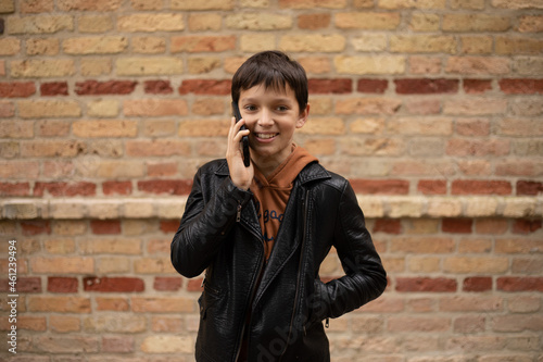 hoto of teenage boy aged 11-12 is talking on smartphone against background of brick wall in the city, smile on his face