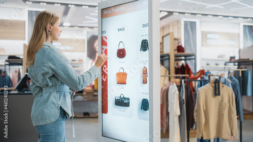Beautiful Female Customer Using Floor-Standing LCD Touch Display while Shopping in Clothing Store. She is Choosing Stylish Bags, Picking Different Designs from Collection. People in Fashionable Shop.