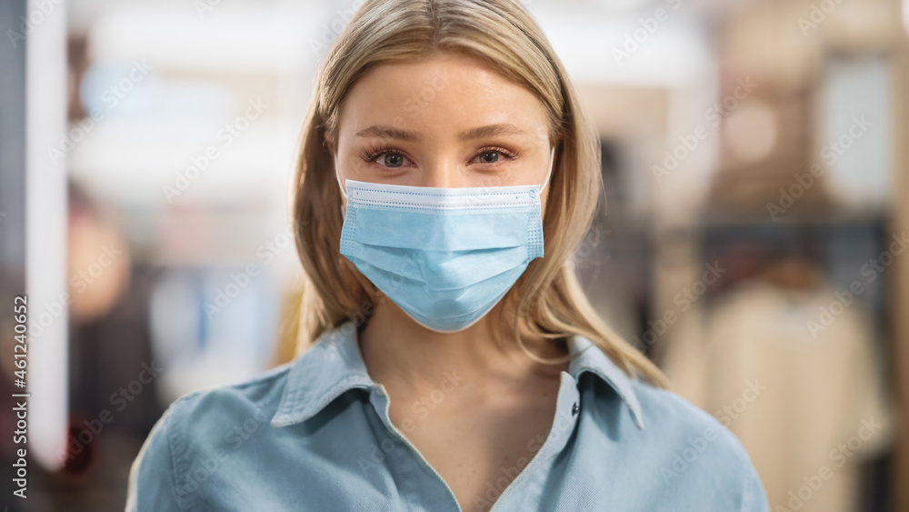 Portrait of a Beautiful Young Woman with Blonde Hair and Green Eyes Wearing Protective Medical Face Mask. Female Practicing Social Distancing, Quarantine. Blurred Clothing Store Background. Portrait of a Beautiful Young Woman with Blonde Hair and Green Eyes Wearing Protective Medical Face Mask. Female Practicing Social Distancing, Quarantine. Blurred Clothing Store Background.