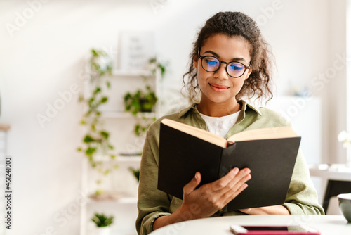 Young black woman in eyeglasses reading book while sitting at cafe