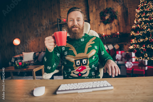 Photo of positive ginger man sit desk hold mulled vine cup watch movie wear sweater decorated office indoors photo