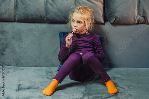 A blonde-haired girl in purple thermal underwear sits brooding on the gray couch in the living room. In her hand is a round lollipop on a stick.