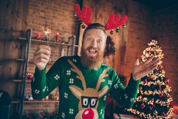 Photo of cheerful guy hold bengal fire stick enjoy atmosphere wear reindeer headband jumper decorated home indoors