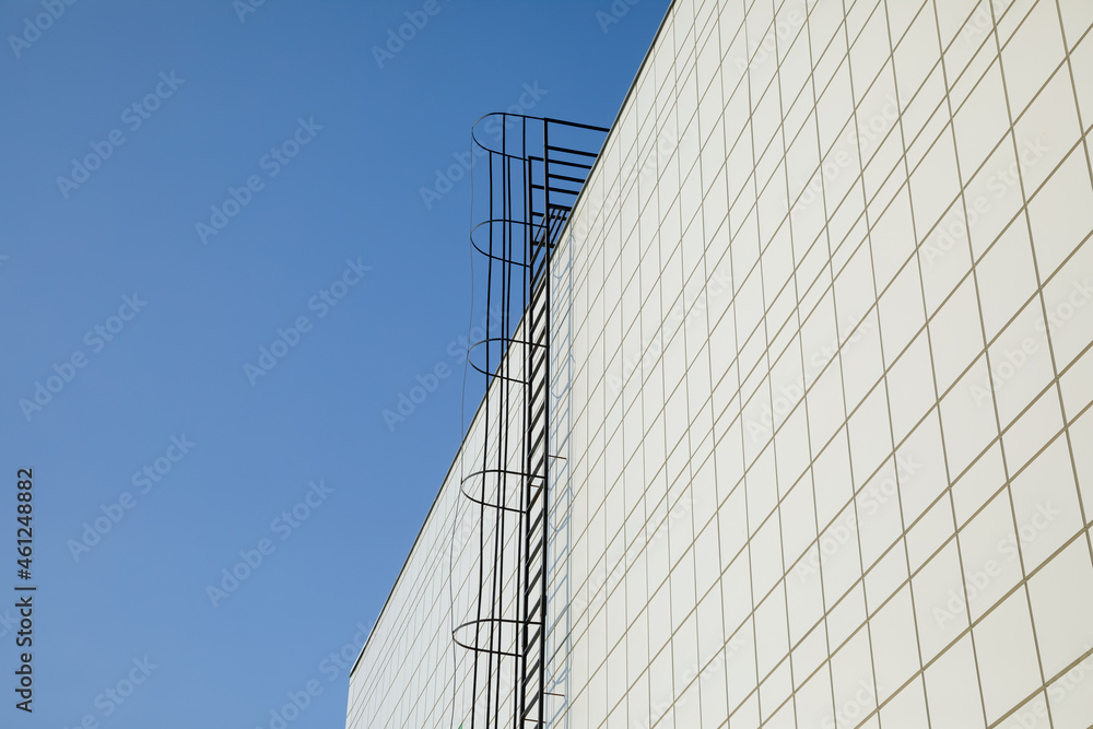 Long metal staircase on the gray modern facade of an industrial building, warehouse or shopping center against the blue sky. Symbol of the stairway to heaven Long metal staircase on the gray modern facade of an industrial building, warehouse or shopping center against the blue sky. Symbol of the stairway to heaven