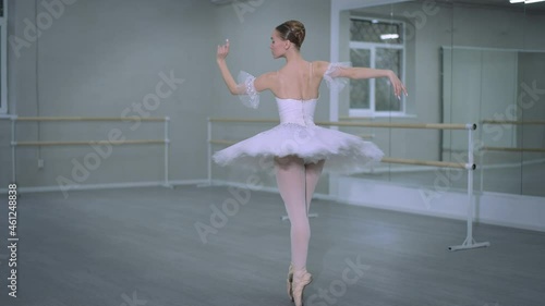 Wide shot graceful ballerina in white tutu spinning on tiptoes in dance studio indoors. Slim Caucasian young woman in pointes rehearsing classical ballroom dance steps. Choreography art and talent photo