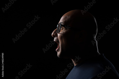 Portrait of a bald man wearing eyeglasses screaming against dark black background.